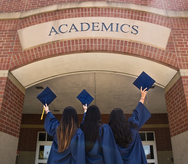 Three graduating students wearing cap and gown standing outside a brick building with the word ‘Academics’ on it
