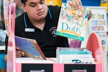 NDIS participant sorting children’s books in a library. He is wearing a black shirt with a logo and a name tag.